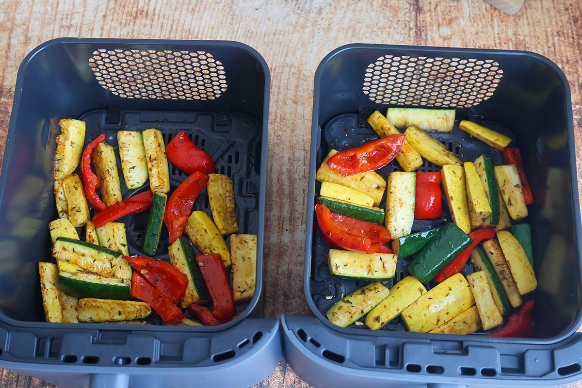 Two air fryer baskets full of sliced squash, zucchini, and bell peppers.