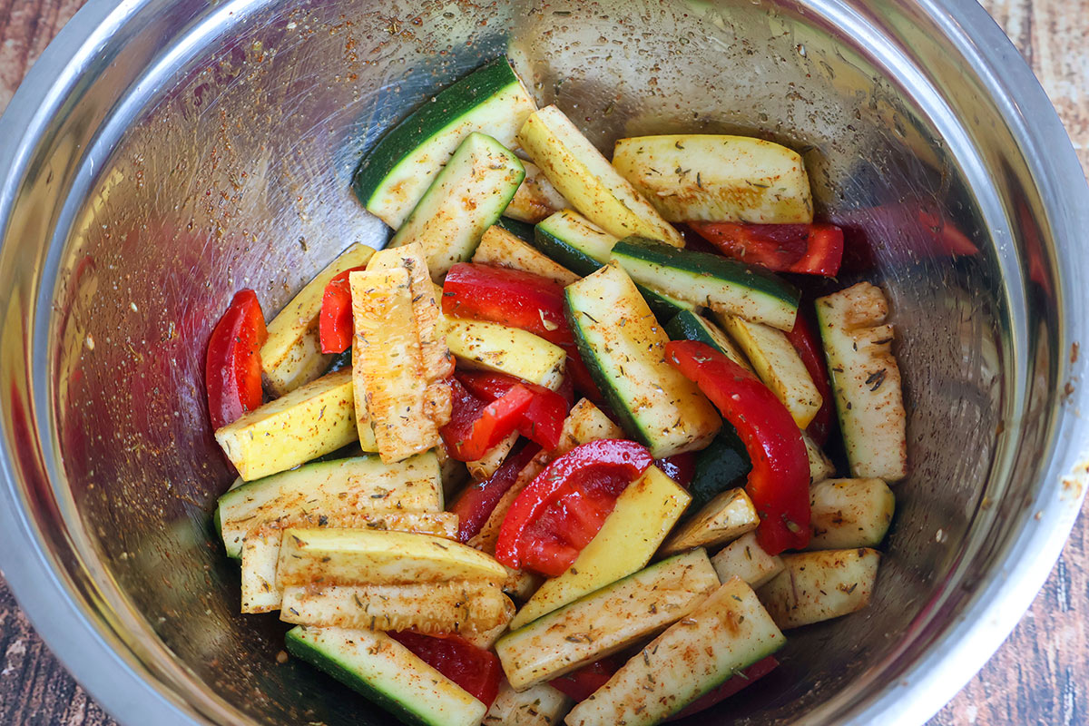 Seasoned, sliced zucchini, squash, and bell pepper in a metal bowl.