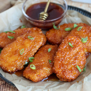 Five hot honey-glazed chicken tenders on a plate with a bowl of hot honey.