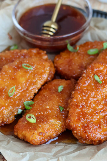 Five hot honey-glazed chicken tenders on a plate with a bowl of hot honey.