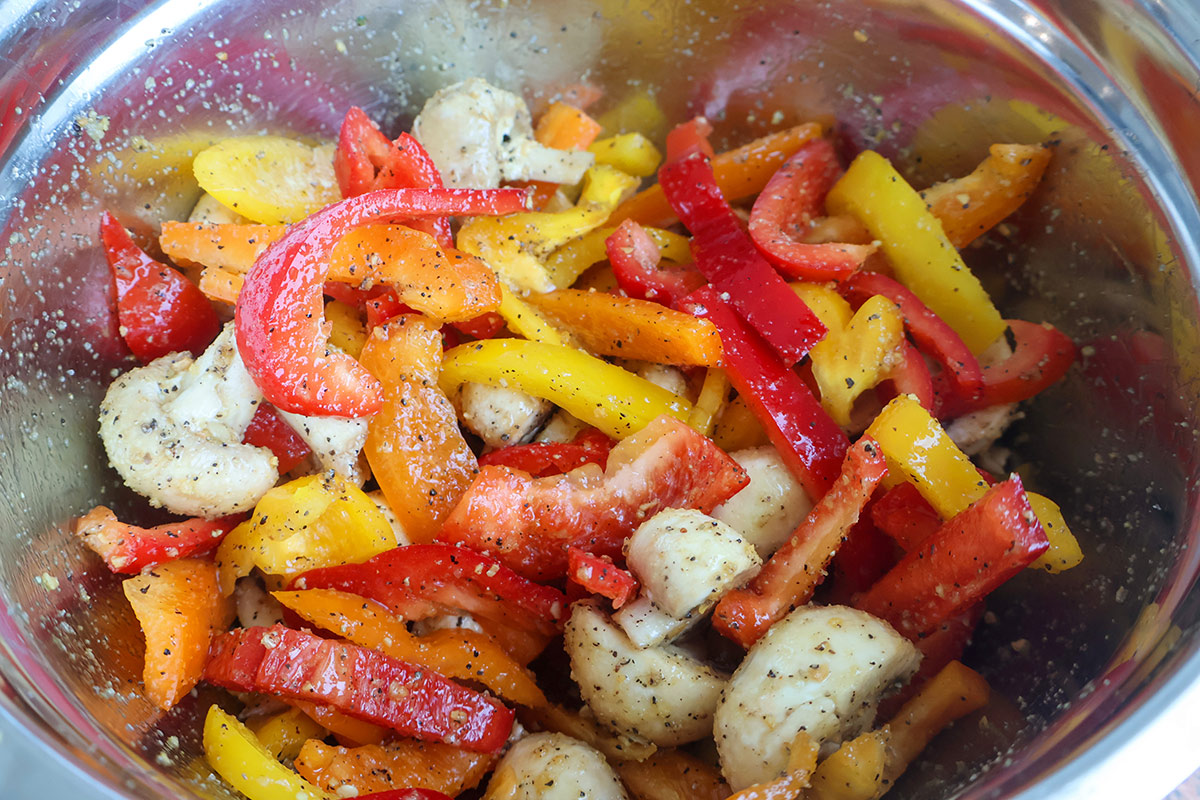 A metal mixing bowl full of seasoned sliced peppers and mushrooms.