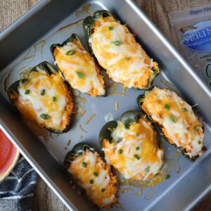 Six stuffed poblano peppers topped with cheese in a baking pan.