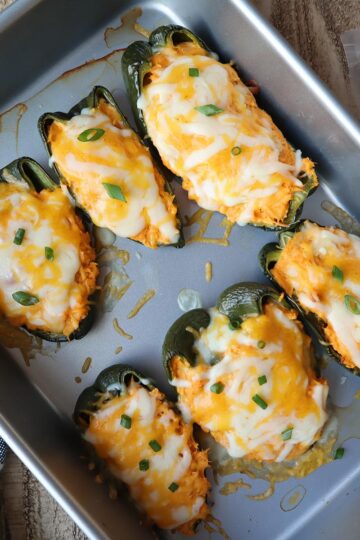 Six stuffed poblano peppers topped with cheese in a baking pan.