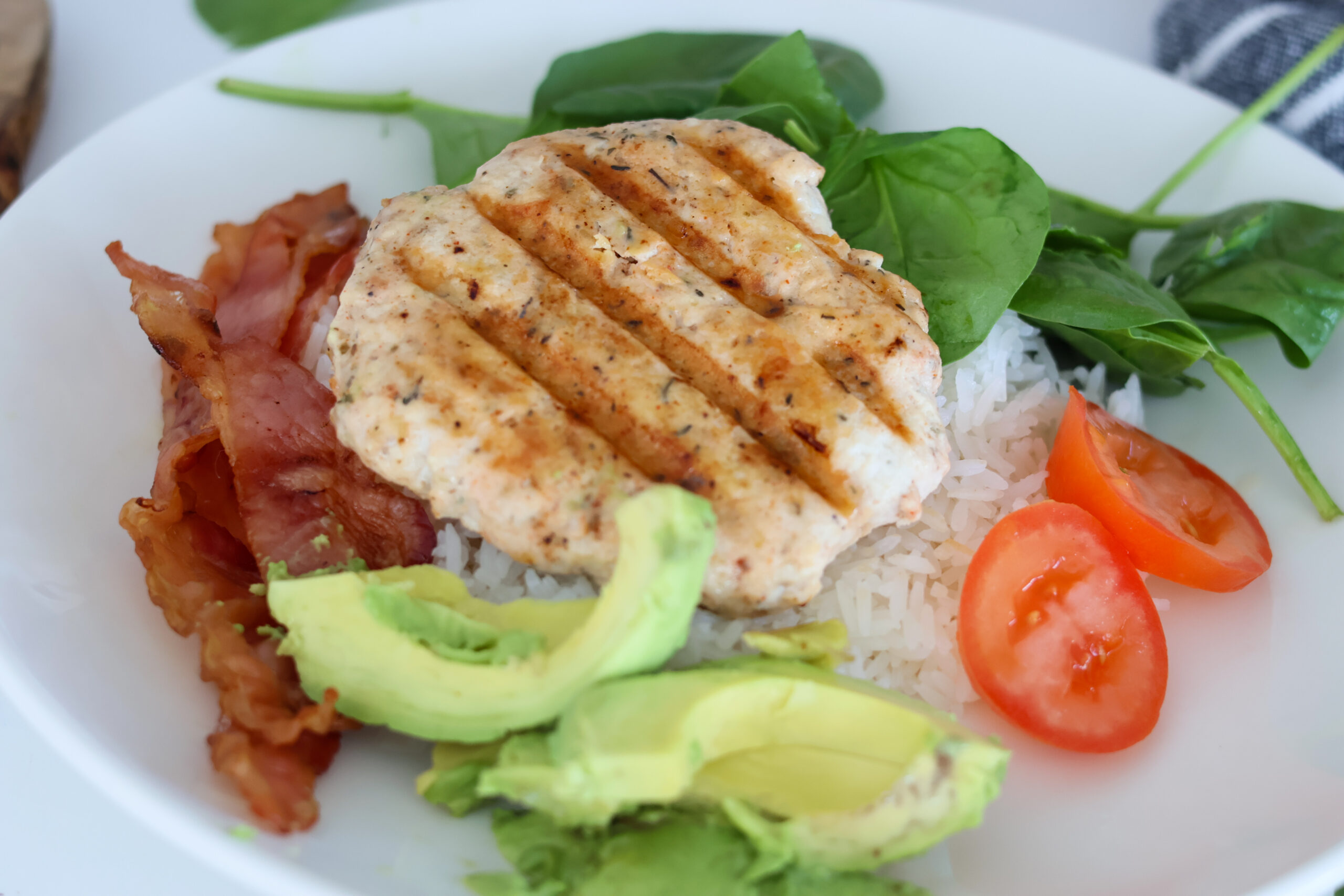 A grilled chicken burger on a plate of rice with bacon and avocado and tomato.