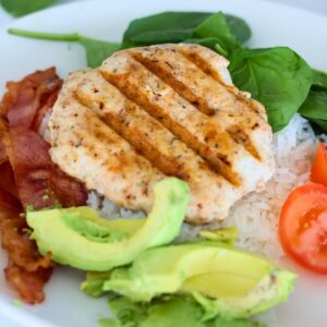A chicken burger with avocado and bacon on a plate of rice.