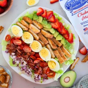 A plate of strawberry cobb salad surrounded by eggs, strawberries and avocado.
