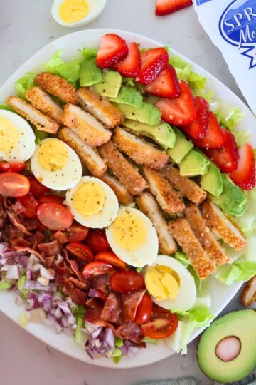 A plate of strawberry cobb salad surrounded by eggs, strawberries and avocado.